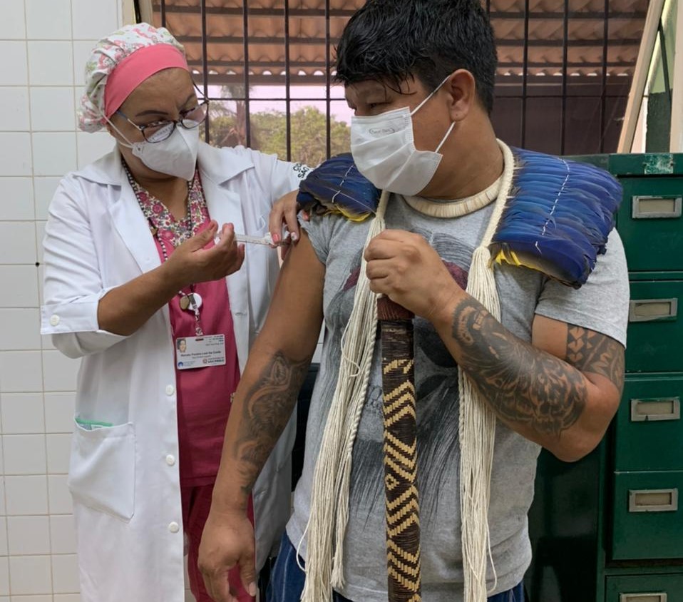 De touca rosa e avental branco, agente de saúde aplica vacina no braço direito do líder indígena Pedro Delane, os dois estão em pé e ele usa camiseta branca e um colar de palha azul nos ombros. A parede da sala é de azulejos brancos e há um armário de metal verde escuro